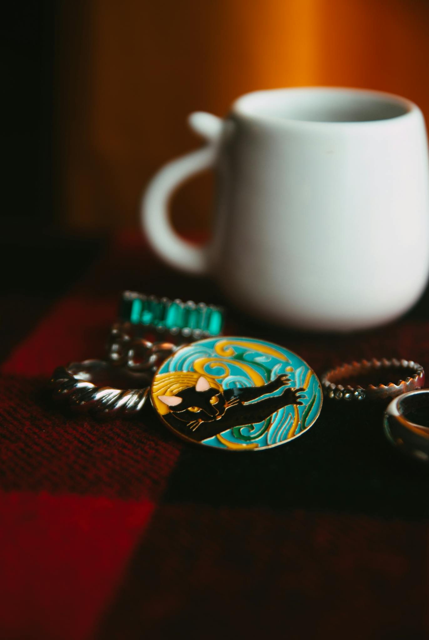 Close-up of artistic jewelry and a white mug on a checkered surface in warm light.